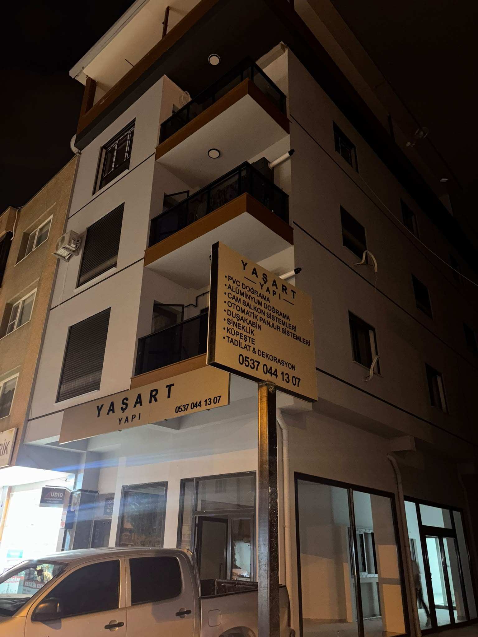 YAŞART YAPI dükkânı gece: köşe binada ışıklı tabela ve hizmet listeli direk tabelası