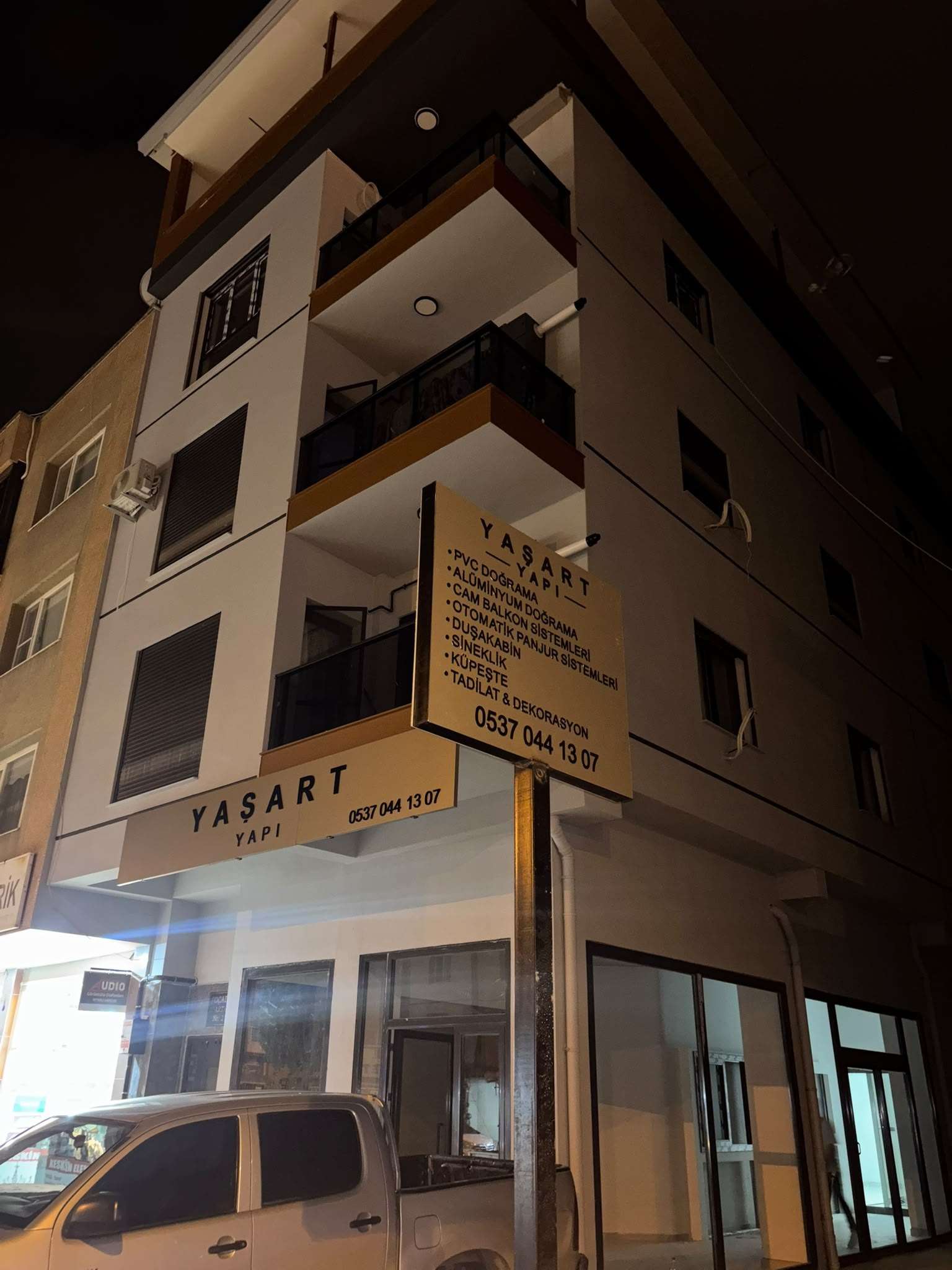 YAŞART YAPI dükkânı gece: köşe binada ışıklı tabela ve hizmet listeli direk tabelası